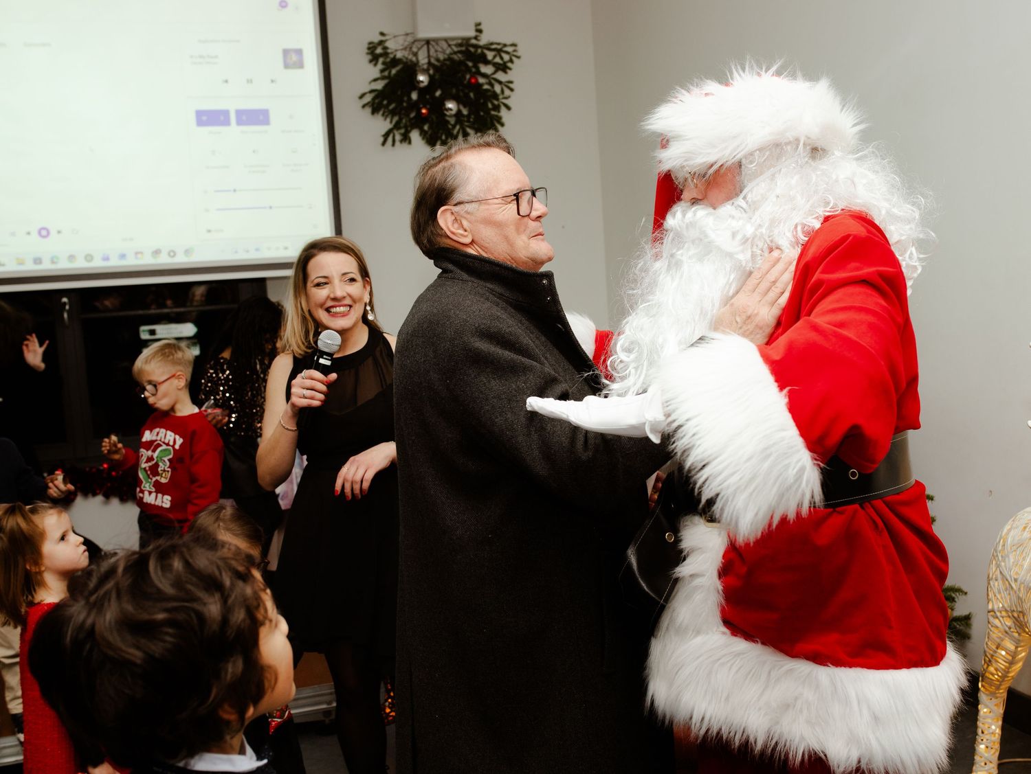 Man greeting Santa Claus while woman holds microphone at a Christmas party event.