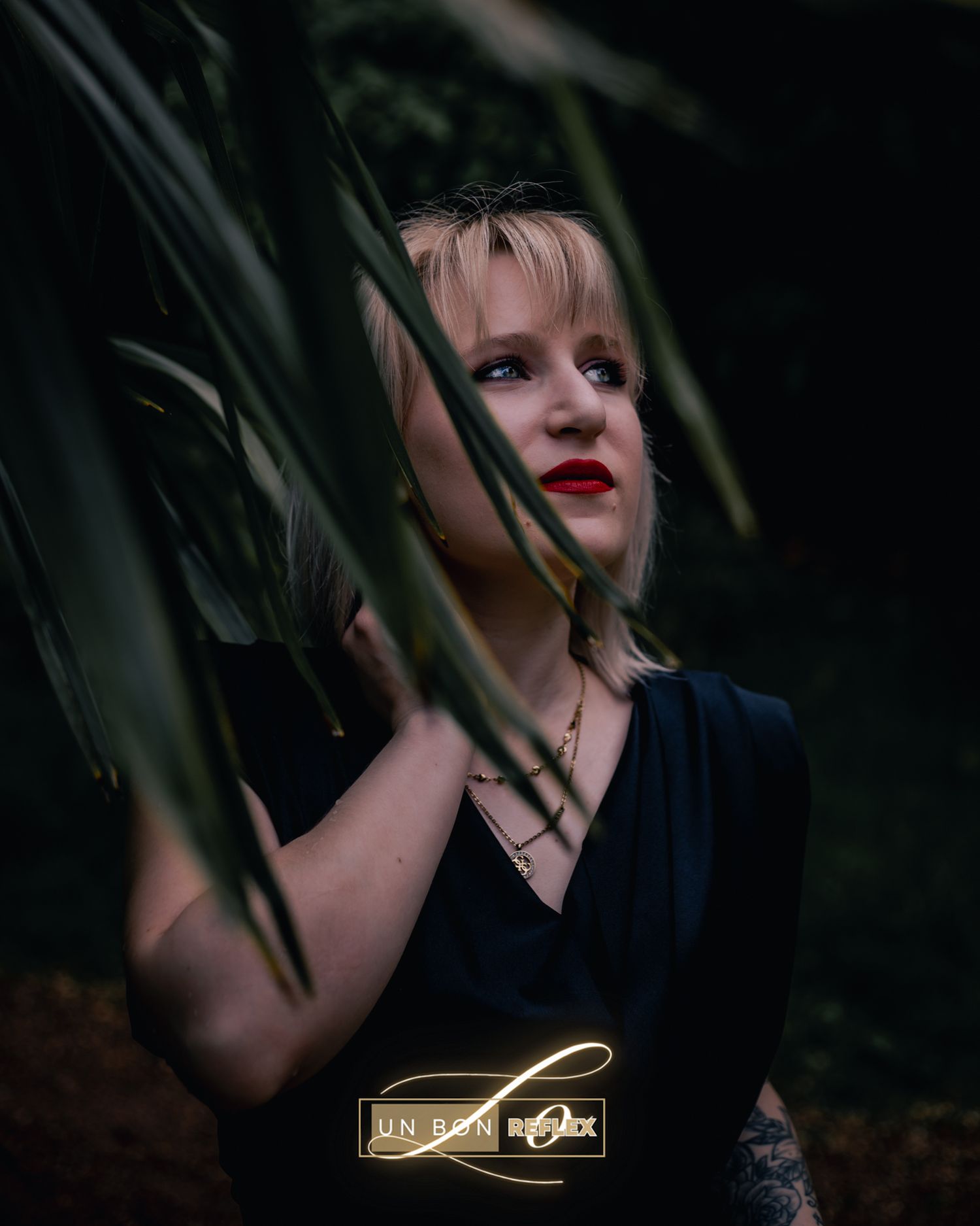 Blonde woman with red lips partially hidden behind tropical palm leaves outdoors.