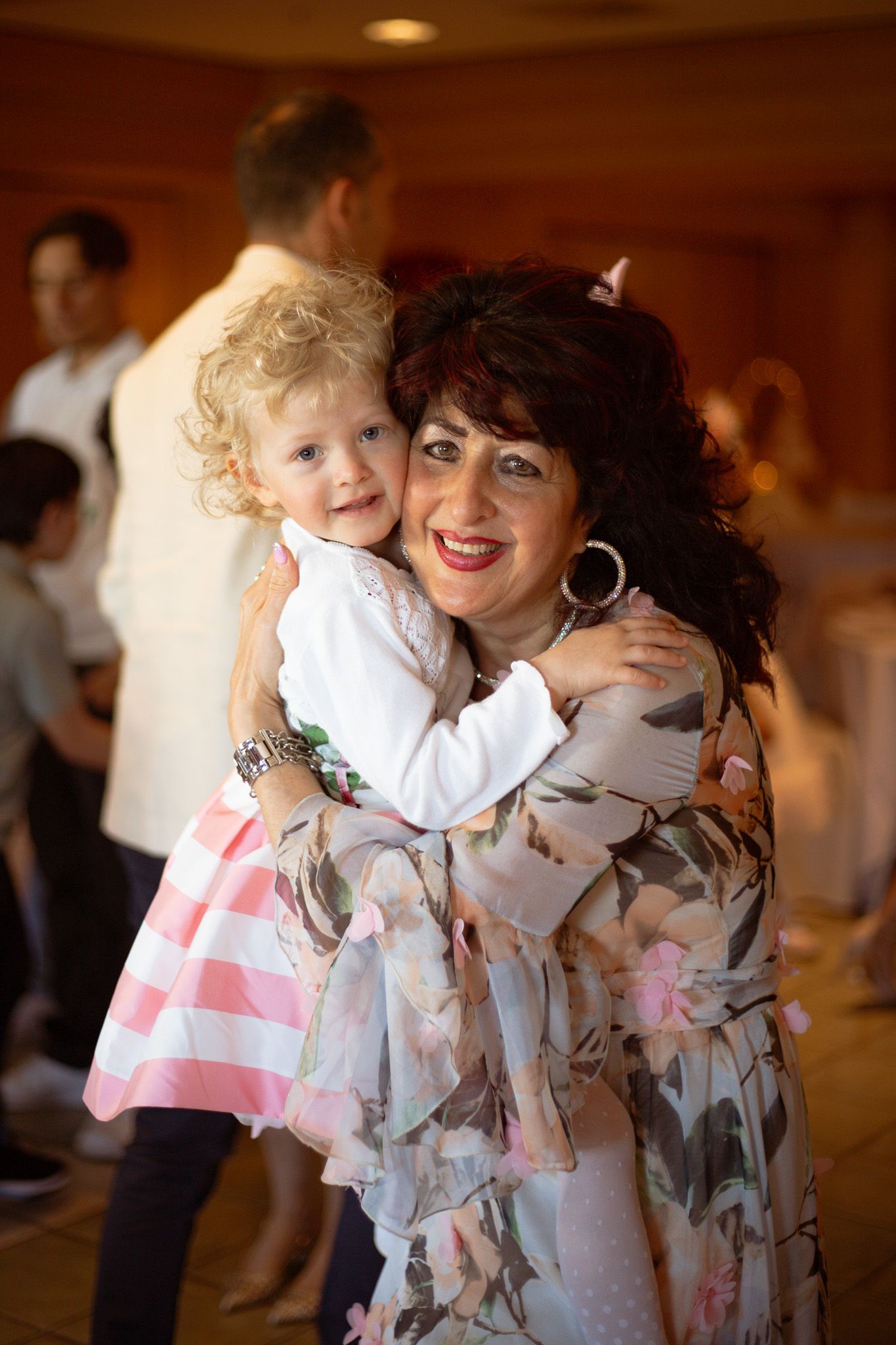 Cheerful grandmother with red lipstick hugging a smiling blonde toddler girl in pink dress at a party.