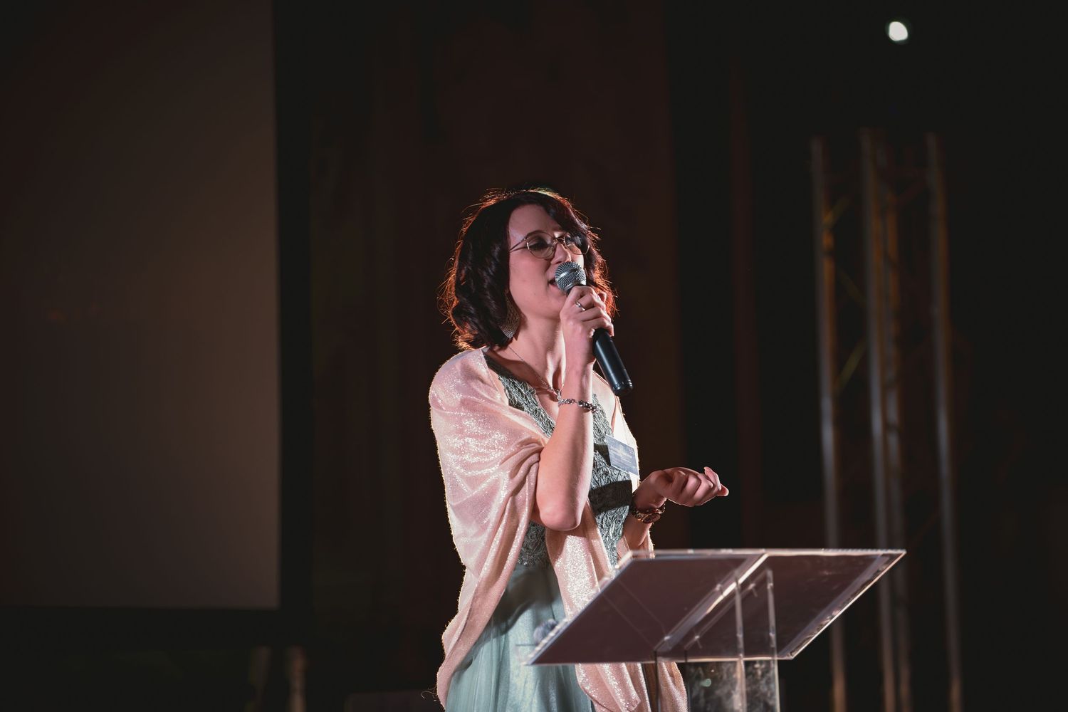 Elegant woman in light blue gown speaking into microphone at podium on stage.