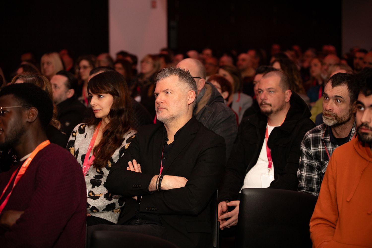 Un public engagé de la conférence regarde une présentation les bras croisés dans un lieu d'événement sombre.