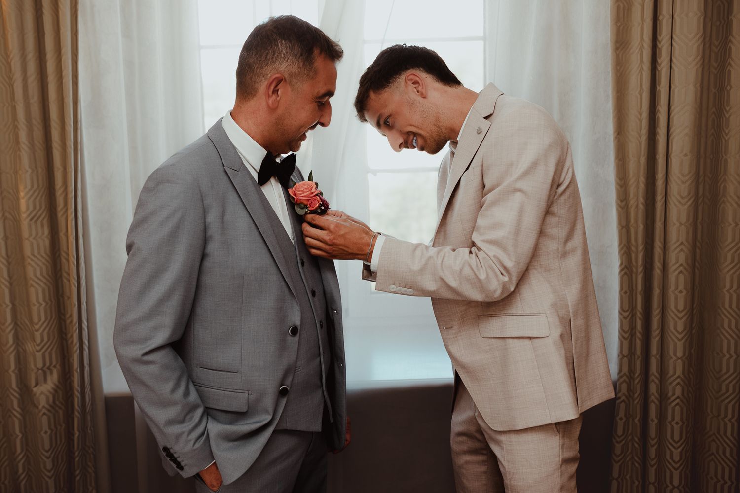 Deux hommes en costume lors d'un mariage, l'un épinglant une boutonnière sur le revers du marié près d'une fenêtre.