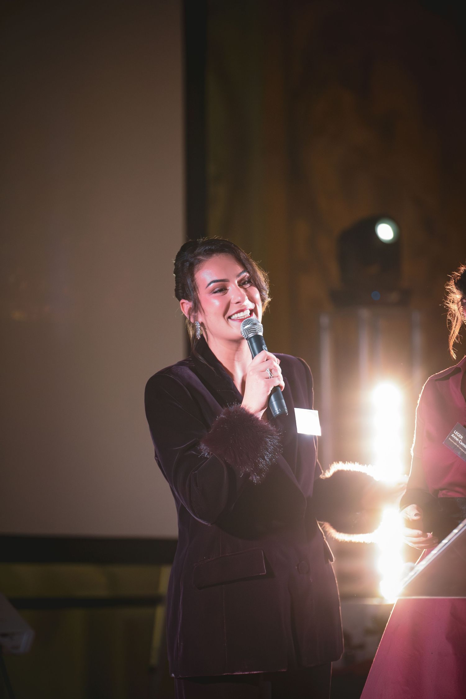Femme souriante en blazer noir parlant dans le microphone sur scène sous un éclairage d'événement lumineux.