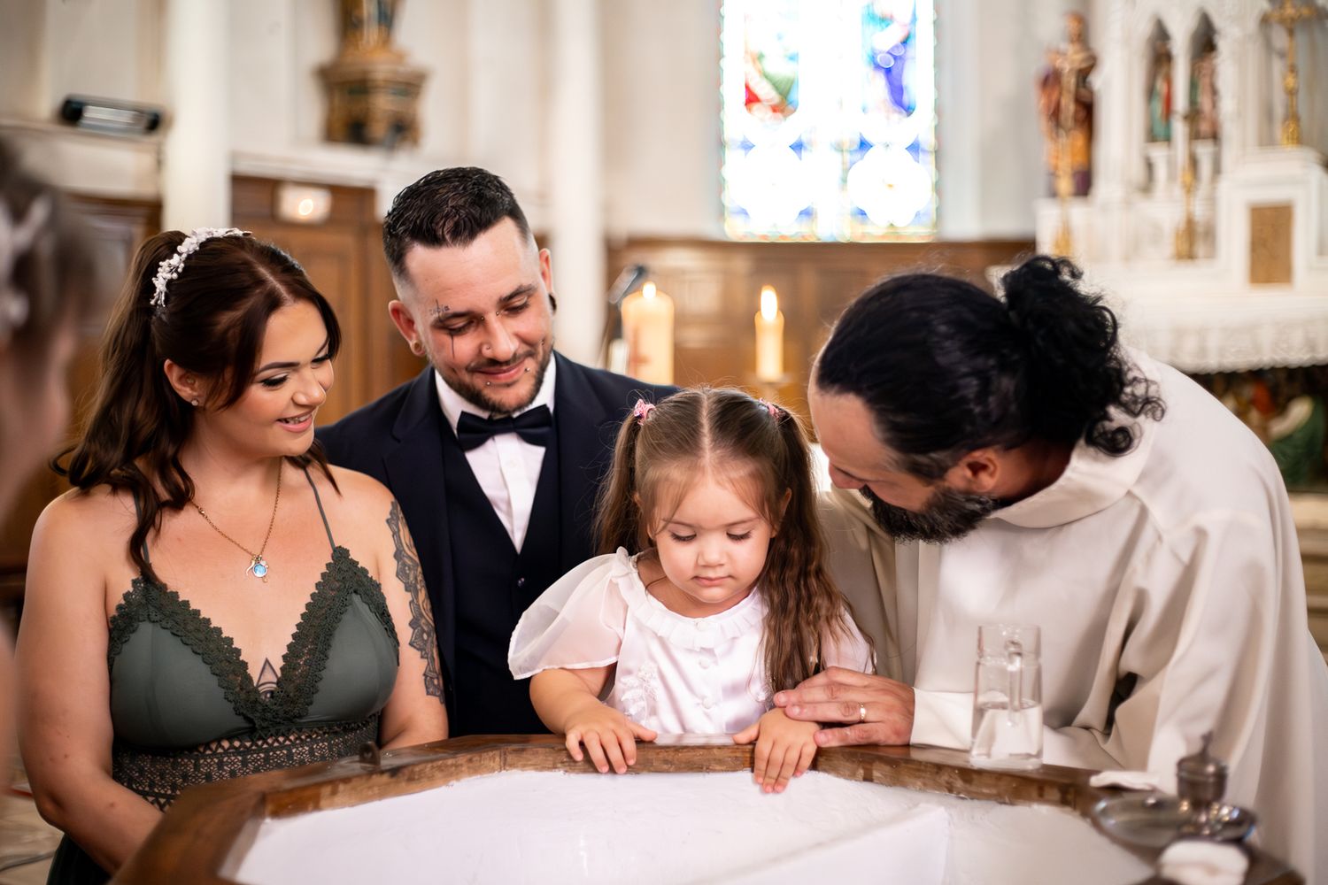 Family gathered around baptismal font with priest during child baptism ceremony in church.