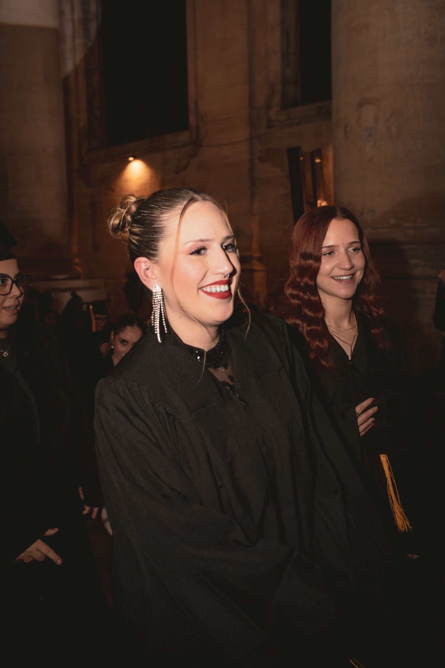 Smiling graduate in black gown with elegant earrings at graduation ceremony.