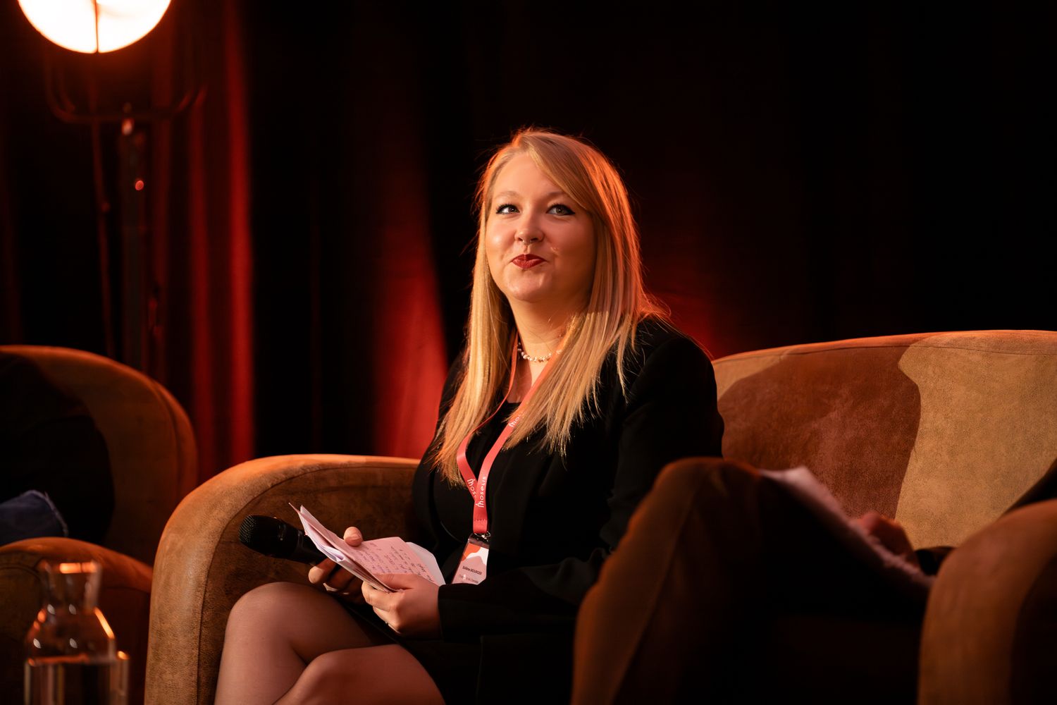 Blonde woman in black dress seated in armchair on stage holding notes at a panel discussion.