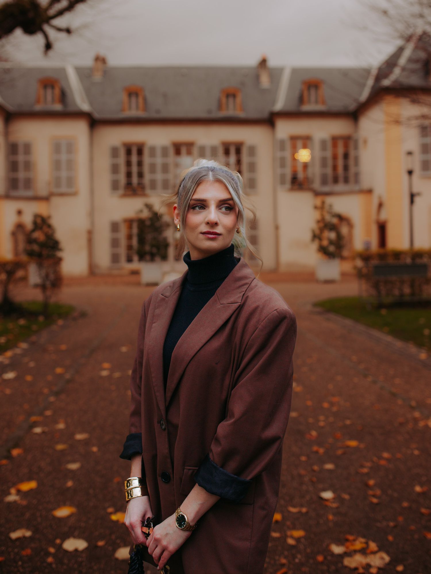 Blonde woman in brown oversized coat standing before elegant French manor at dusk.