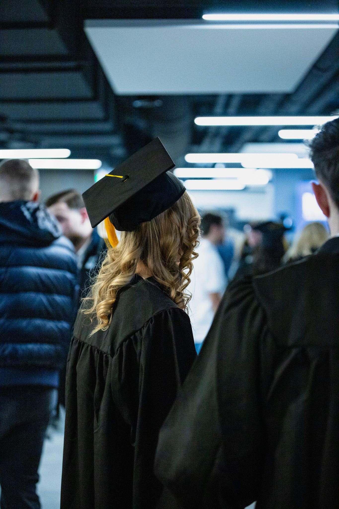 Blonde graduate in black cap and gown seen from behind in hallway.