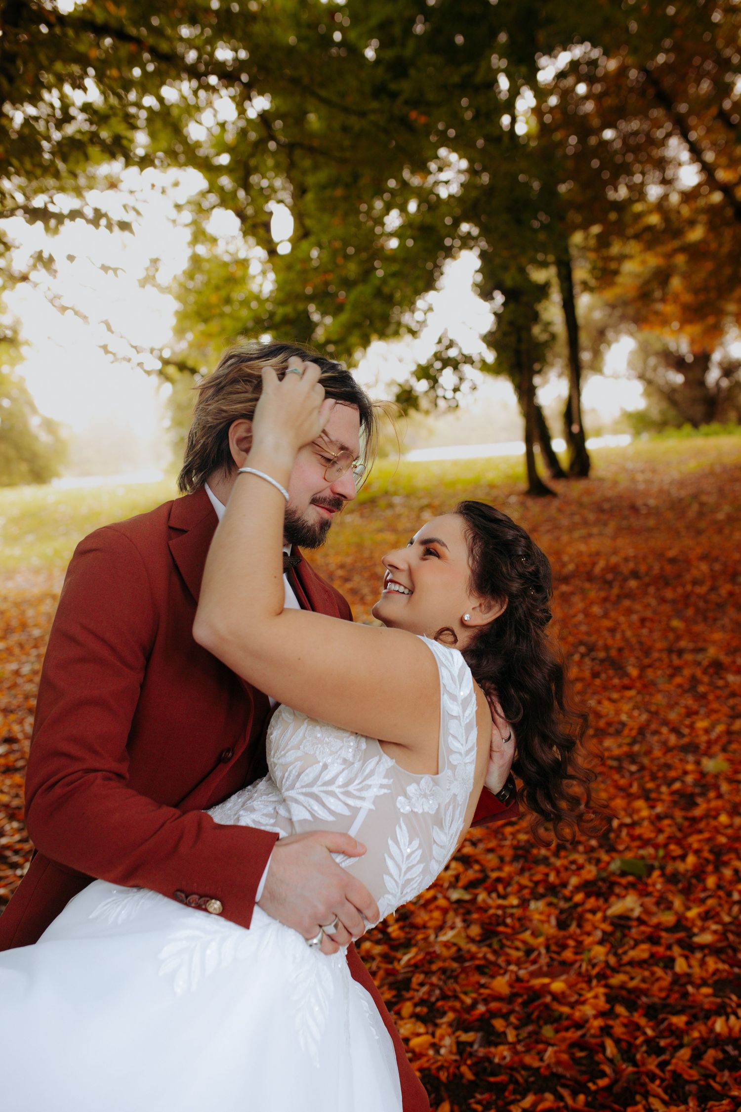 Joyful bride and groom embracing among vibrant autumn fallen leaves in a sunlit park during their wedding.