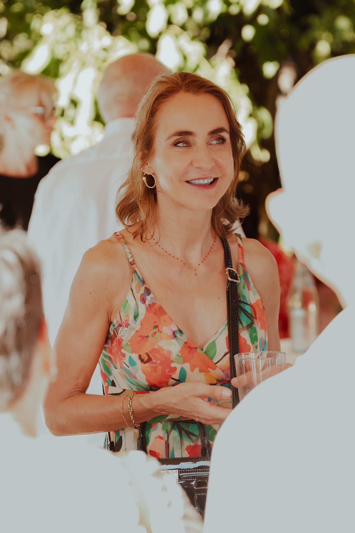 Woman in floral dress smiling at outdoor garden party, holding a drink, wearing gold earrings and necklace.