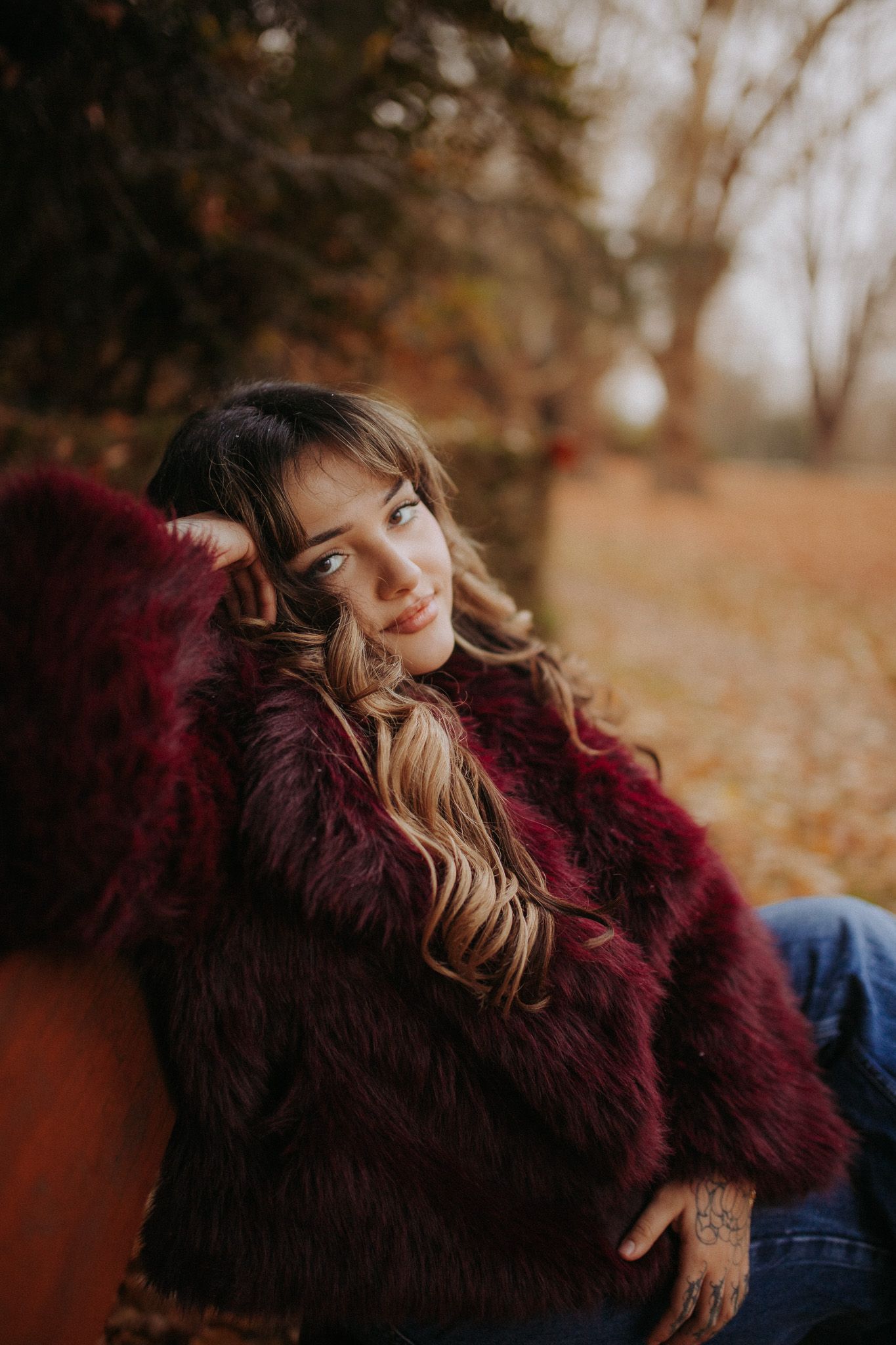 Young woman in dark red faux fur coat sitting outdoors in autumn park setting.