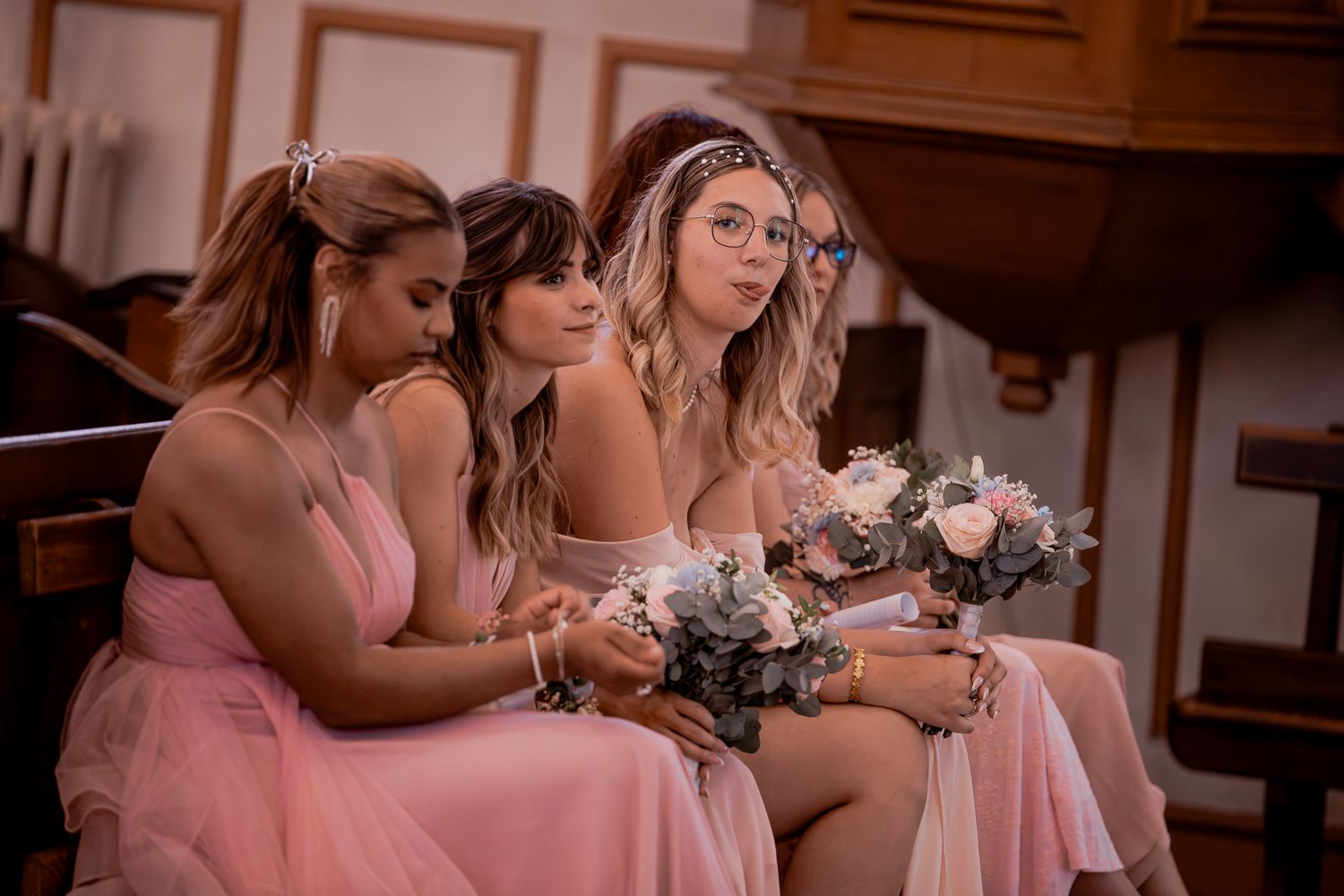 Three bridesmaids in pink dresses holding floral bouquets seated in a church during a wedding ceremony.