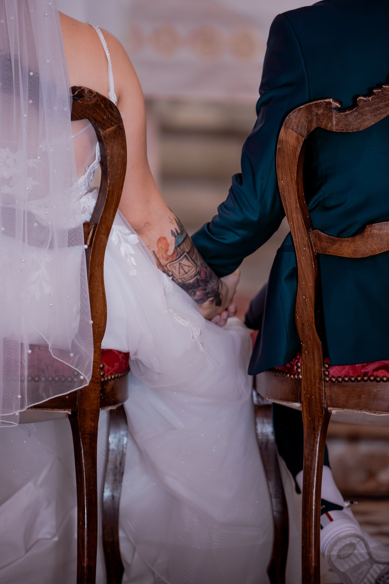 Close-up of tattooed bride and groom holding hands while seated in ornate wooden chairs during wedding ceremony.