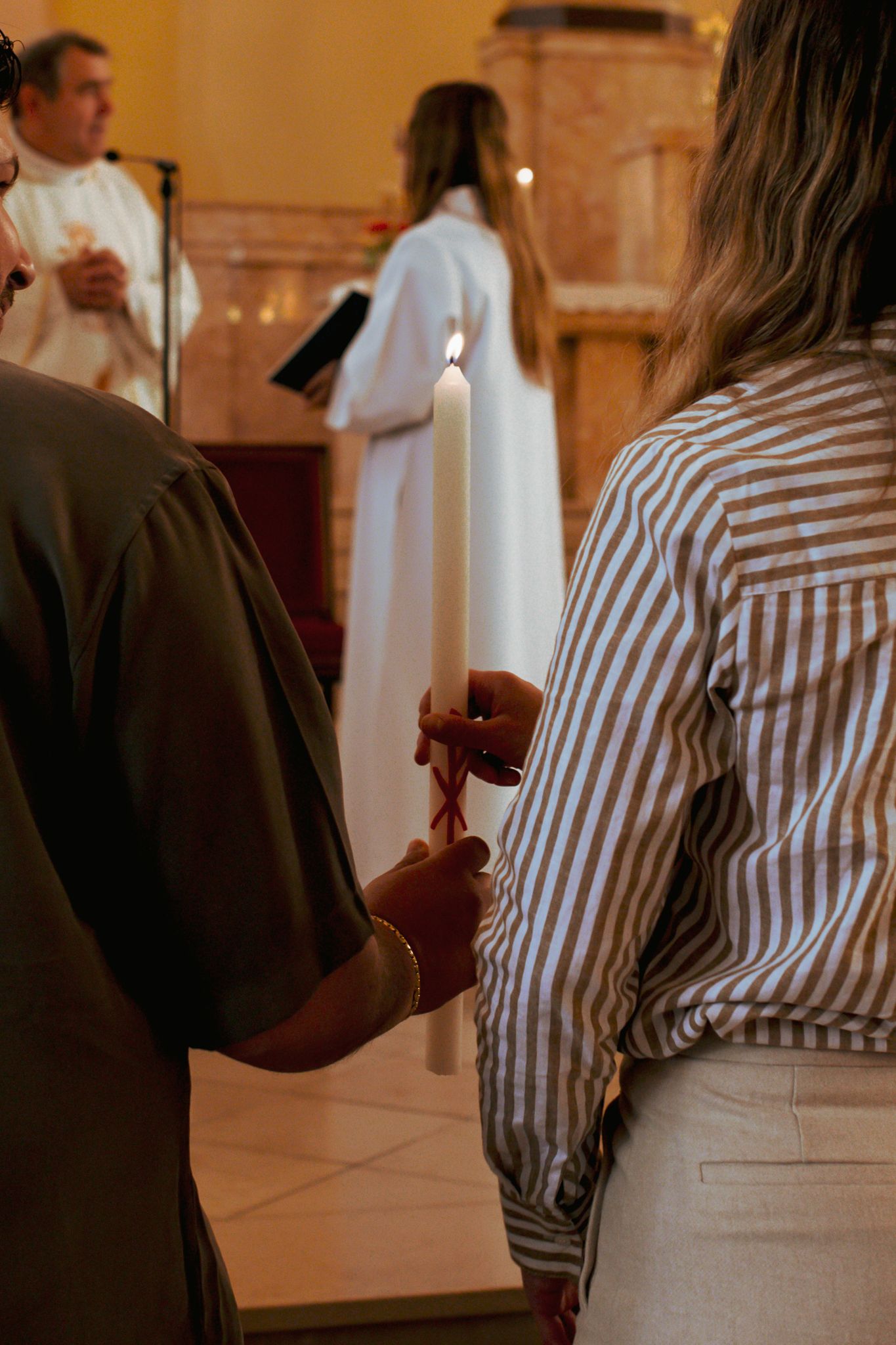 Woman in striped shirt holds a lit white baptism candle during a church ceremony.
