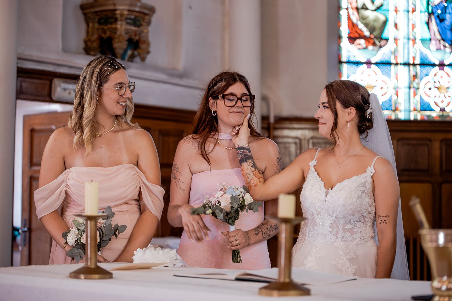 Bride laughing with two bridesmaids in blush off-shoulder dresses near candles and stained glass in a church.
