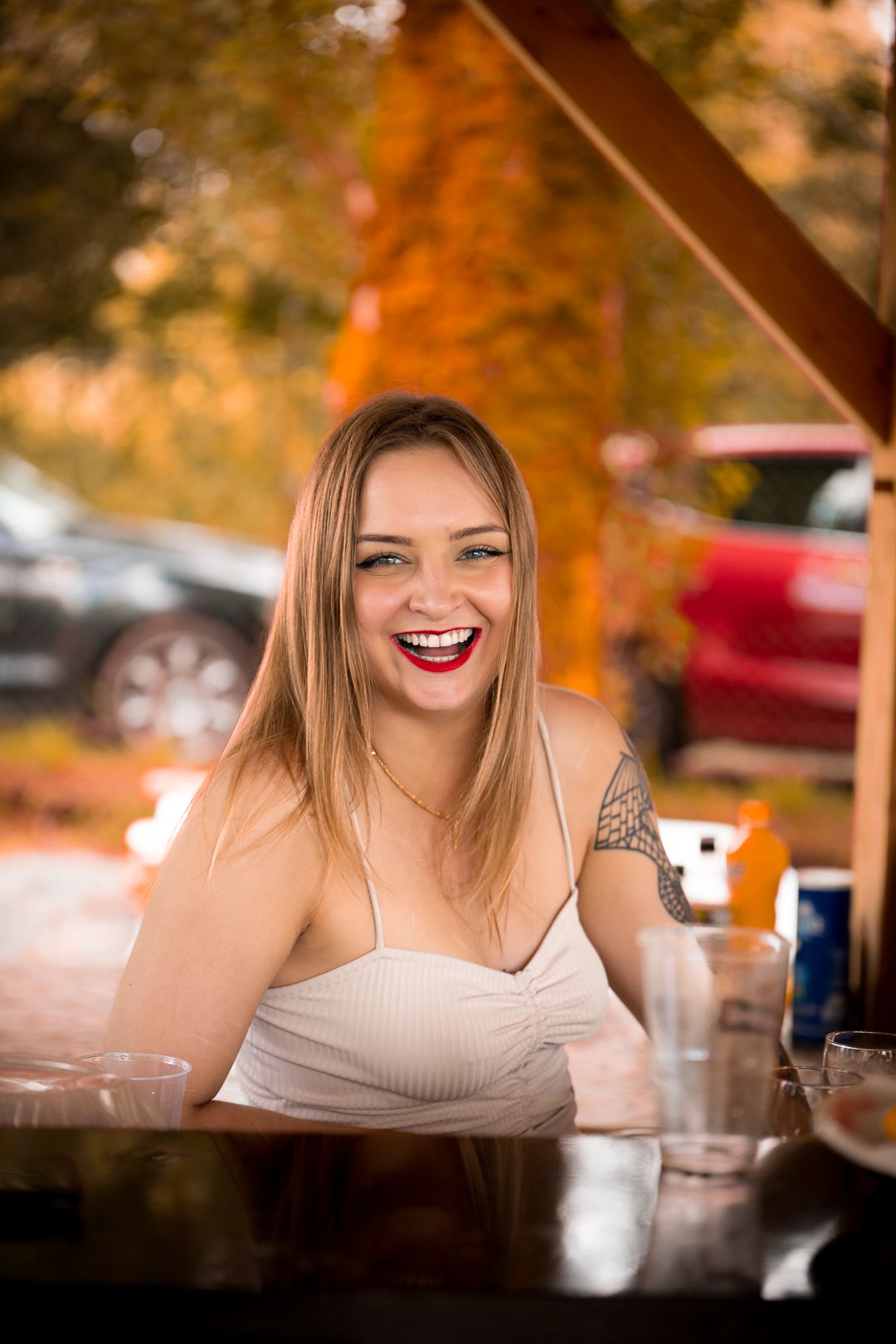 Laughing blonde woman with red lipstick and tattoo seated at an outdoor bar.