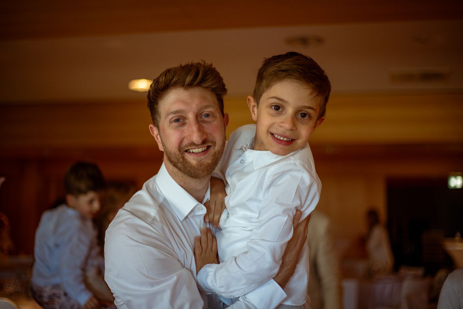 Smiling man in white shirt holds a happy young boy at a warm indoor celebration event.