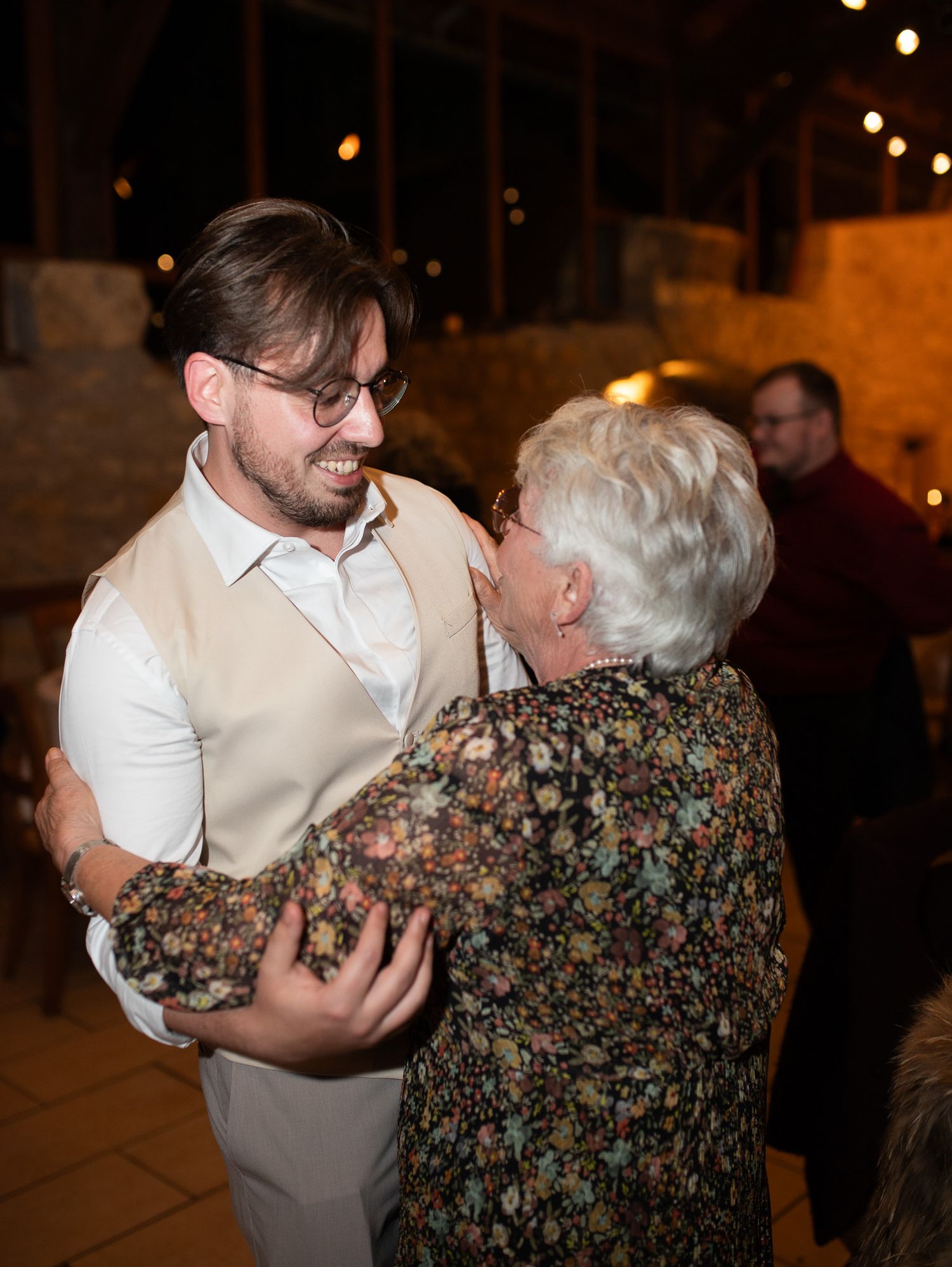 A young man in a vest dances with an elderly woman at a warmly lit evening reception.