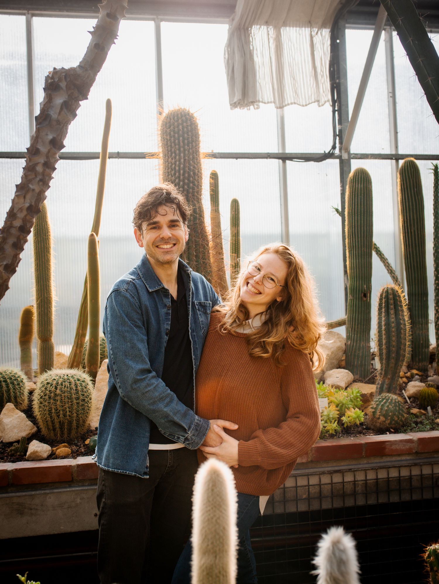 A smiling couple poses in a sunlit greenhouse surrounded by tall cacti, the woman cradling her pregnant belly.