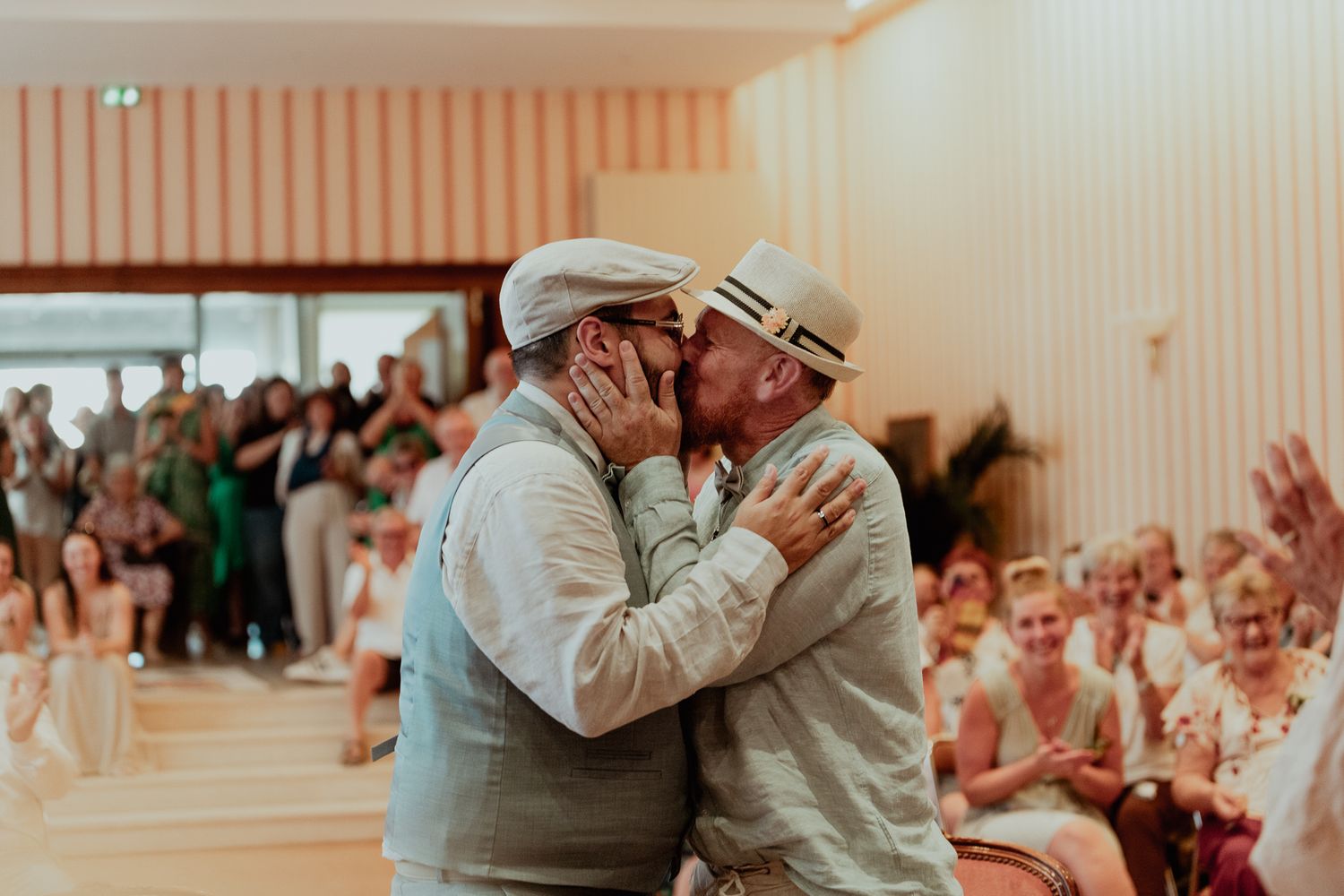 Two grooms sharing a first kiss as newlyweds surrounded by cheering wedding guests.
