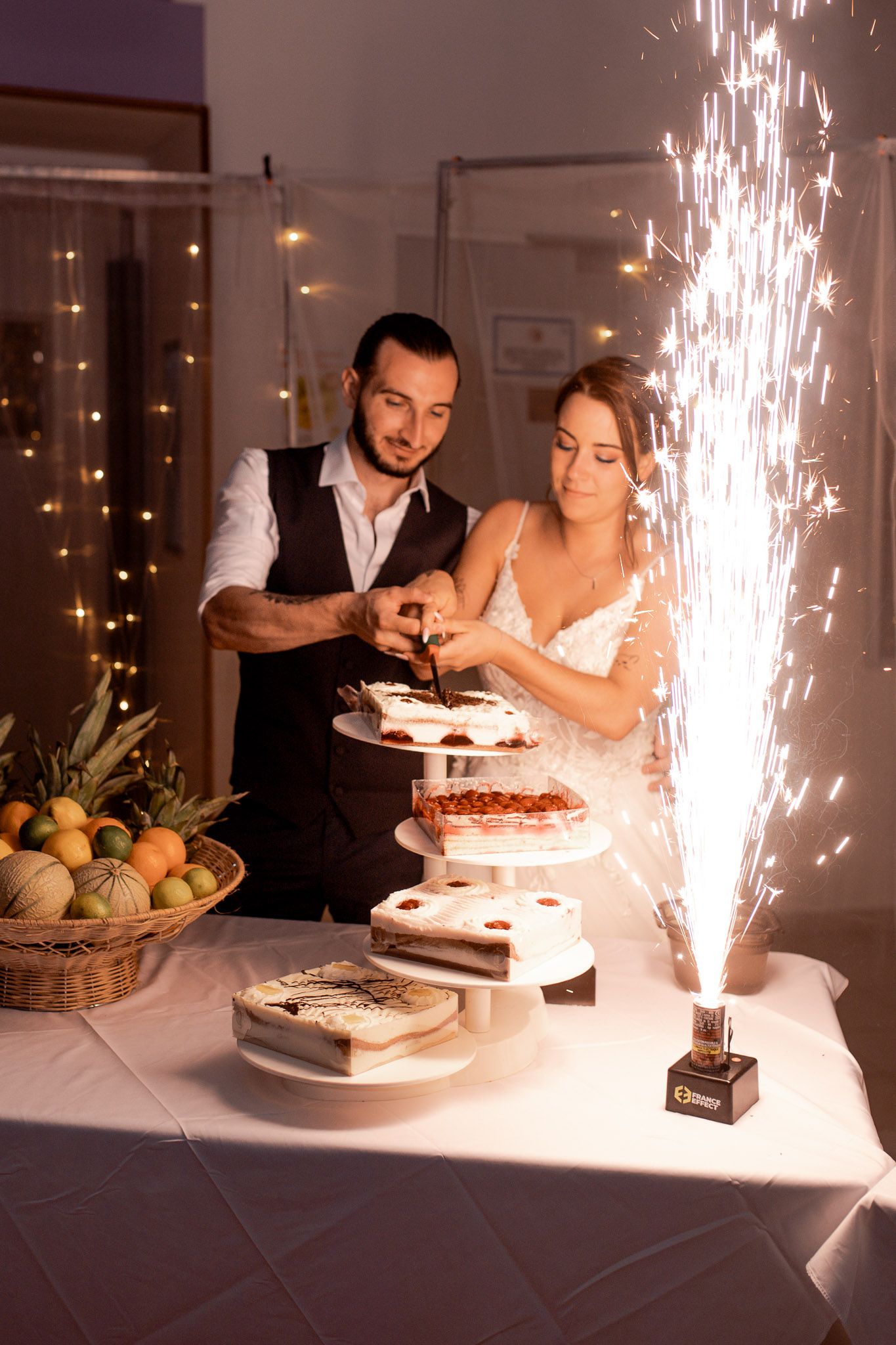 Bride and groom cut their elegant tiered wedding cake beside a dazzling sparkler fountain display.