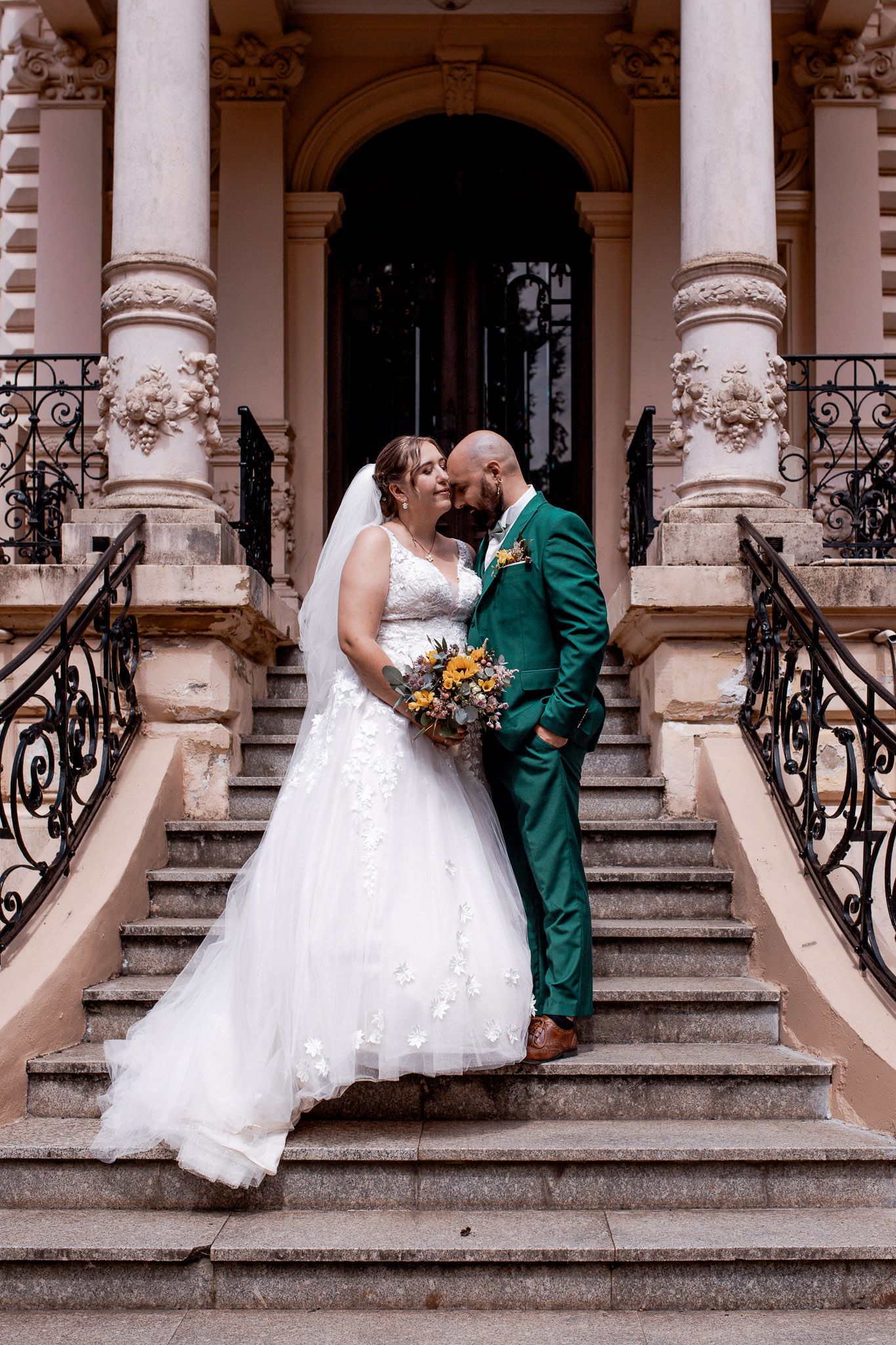 Bride and groom share a kiss on grand ornate staircase, bride holding a yellow bouquet.