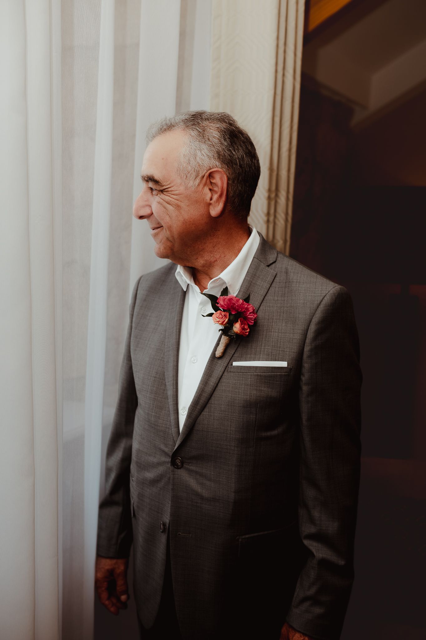 Elegant older man in gray suit with pink boutonniere standing by white curtains at a formal event.