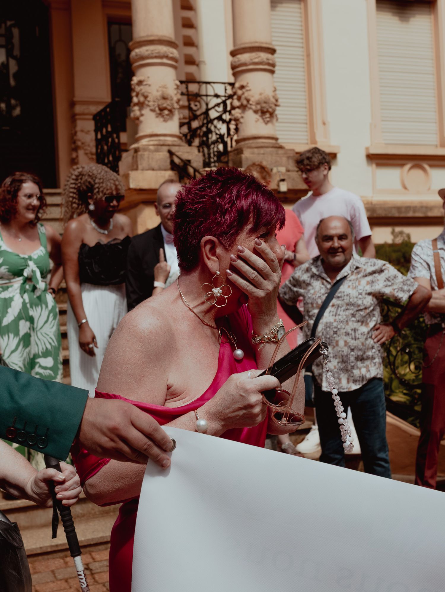 Woman with red hair in pink outfit covers her mouth, surrounded by guests outside an ornate building.
