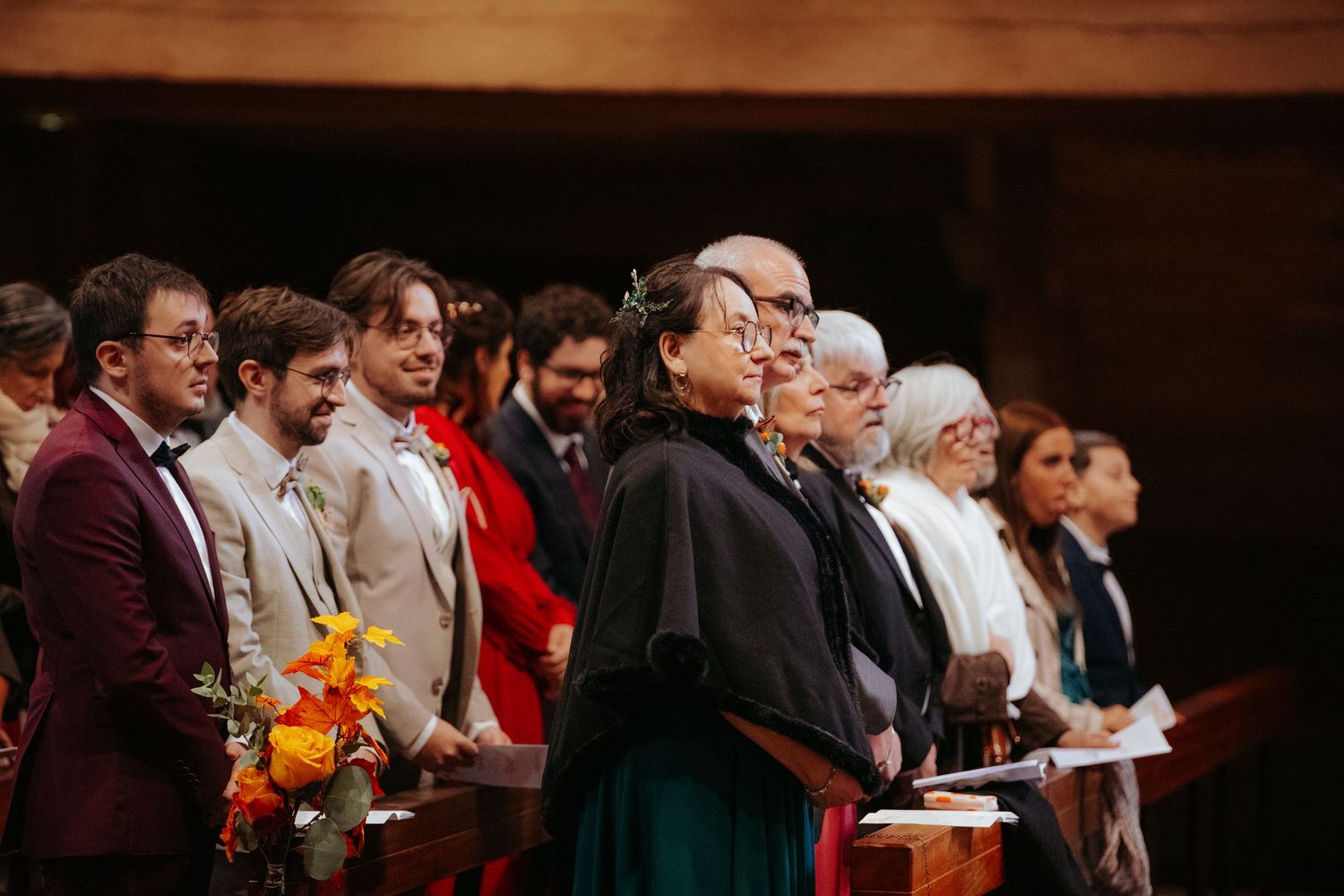 Wedding guests standing in church pews during a ceremony, holding flowers and programs in a warm wooden interior.