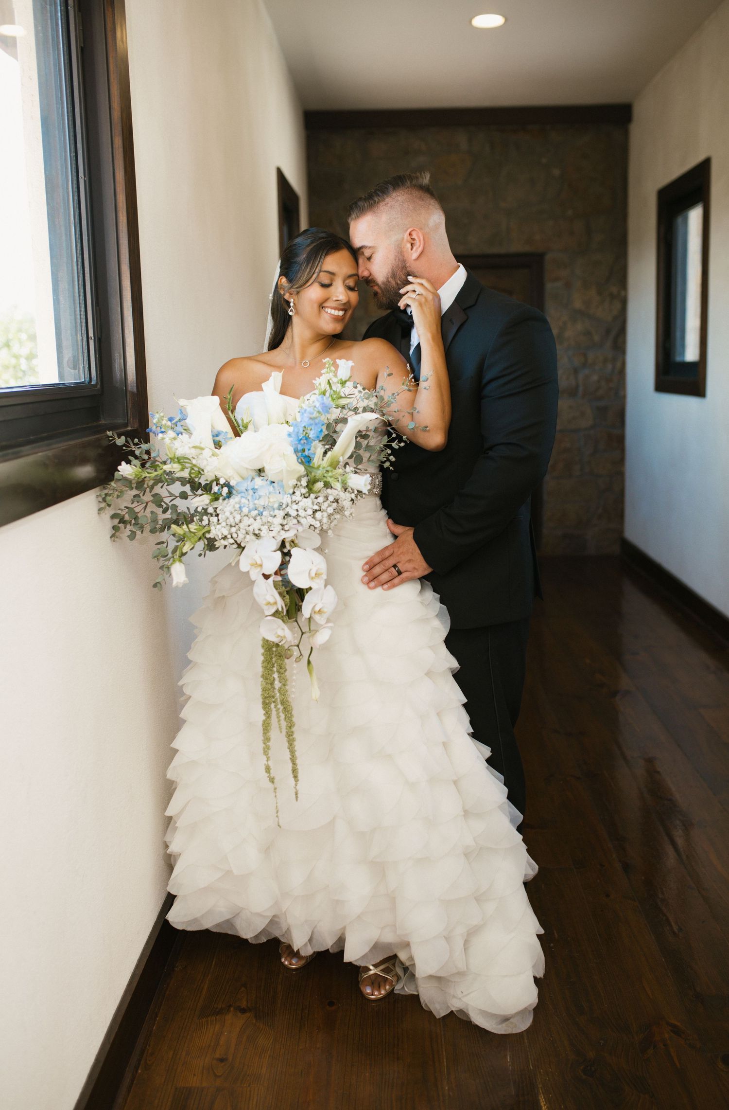 Bride in ruffled white gown holding blue and white bouquet with groom in hallway.