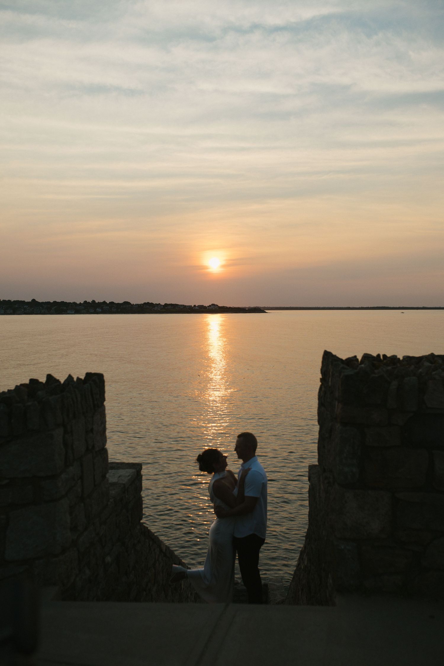Couple silhouetted against a stunning sunset over calm water, framed by stone walls.