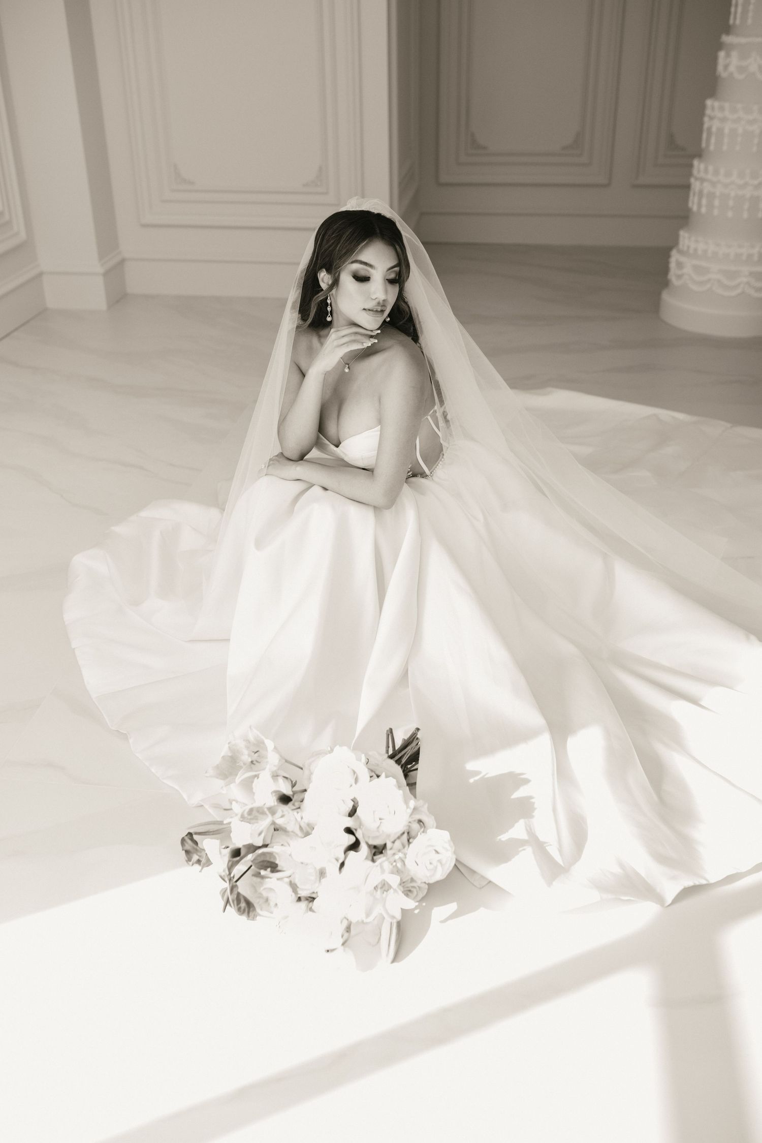 Black and white photo of Asian bride in satin gown with long veil seated near white floral bouquet.