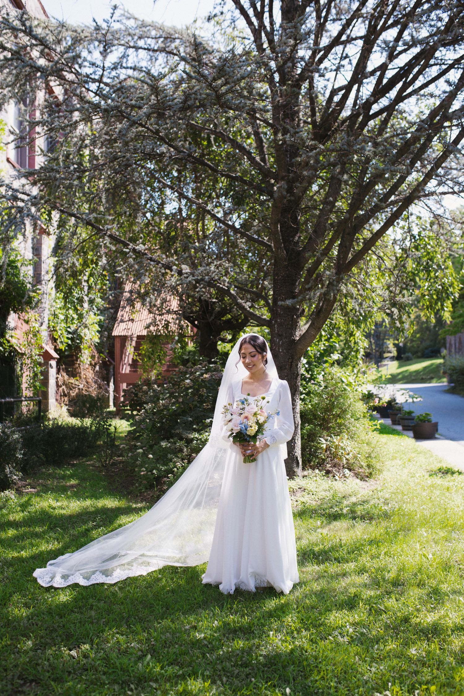 Bride in white gown holds floral bouquet standing on green lawn beneath blooming tree.
