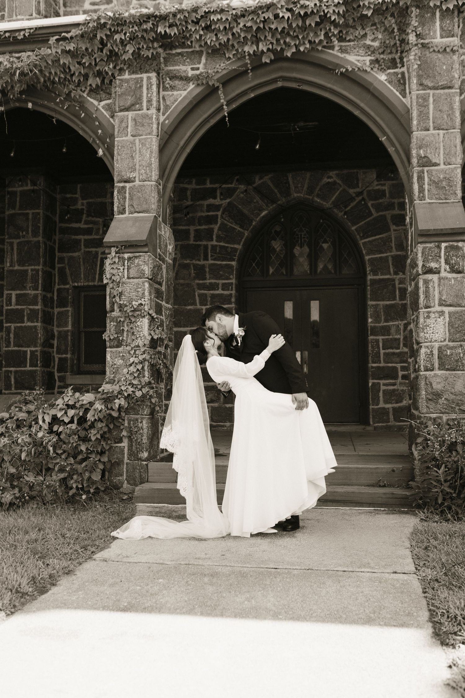 Bride and groom share a romantic dip kiss outside a stone church in black and white photo.