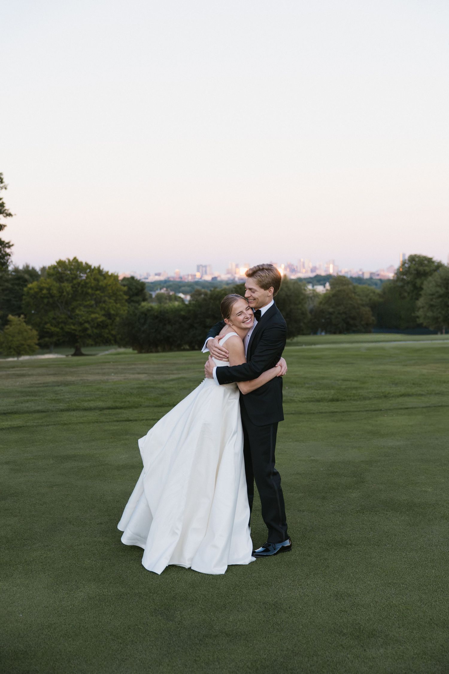 Bride and groom embrace on golf course lawn at dusk with city skyline visible in background.
