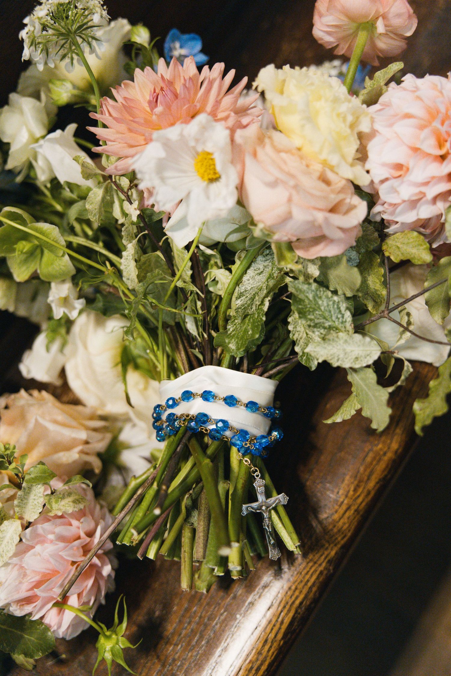 Wedding bouquet of pink roses and dahlias wrapped with blue rosary beads on wooden surface.