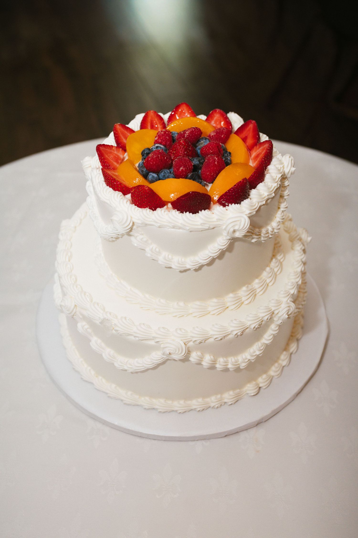 Elegant three-tier white wedding cake topped with fresh strawberries, peaches, and blueberries on a white table.