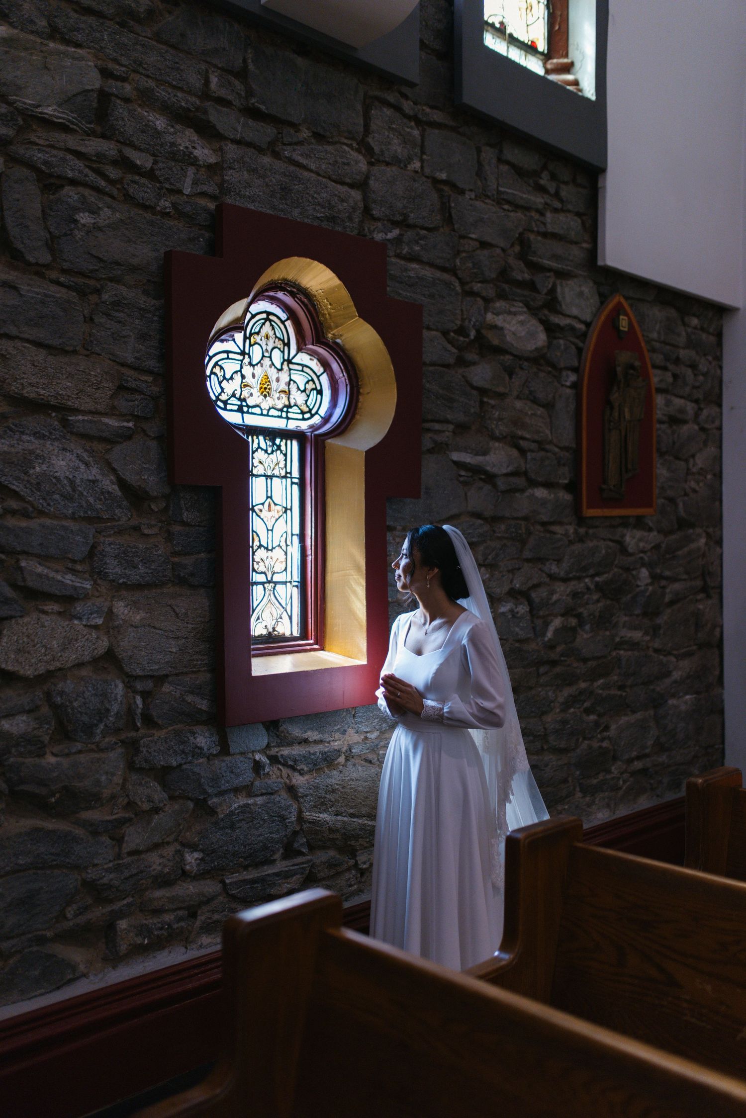 Bride in white gown prays beside stained glass window in stone church interior with soft light.