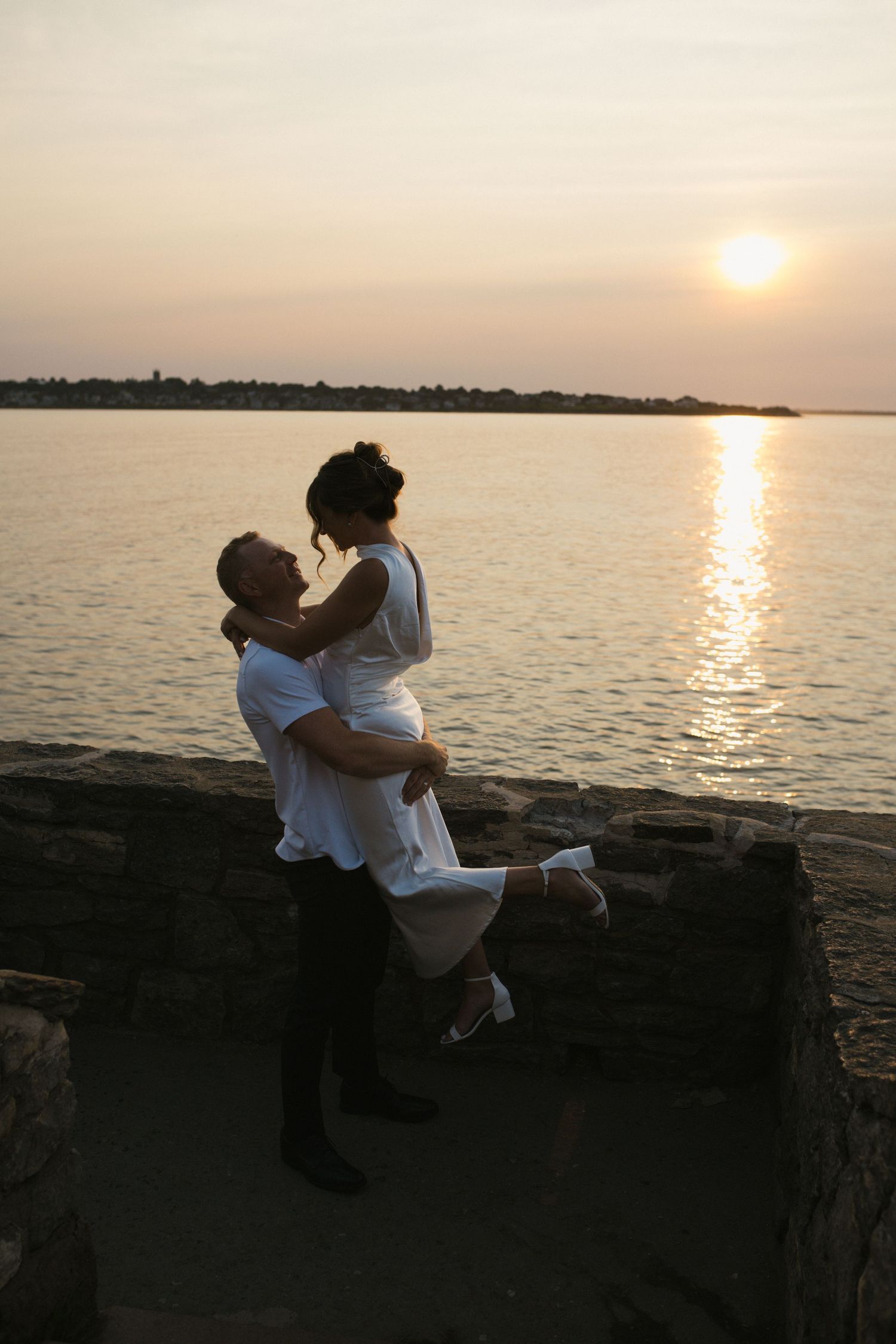 Couple embracing romantically at sunset by the water, man lifting woman in white dress on stone wall.