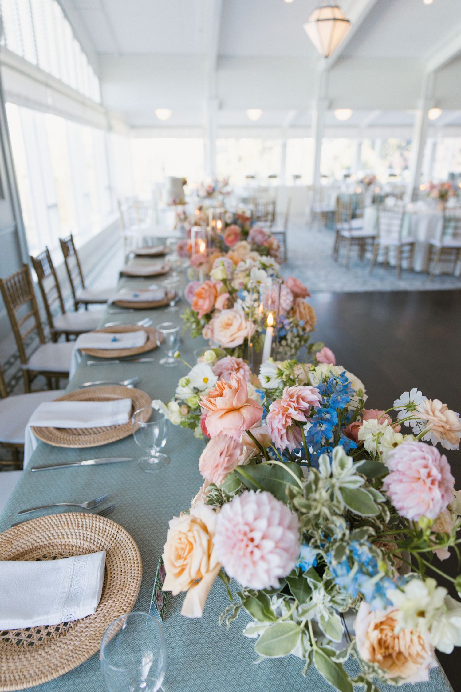 Elegant wedding reception table with pastel floral centerpieces, wicker chargers, and blue tablecloth in bright venue.