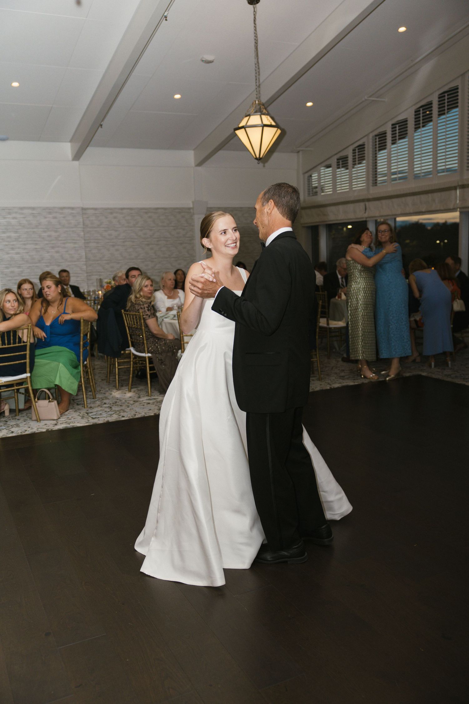 Bride and groom share their first dance in a warmly lit wedding reception hall with guests watching.