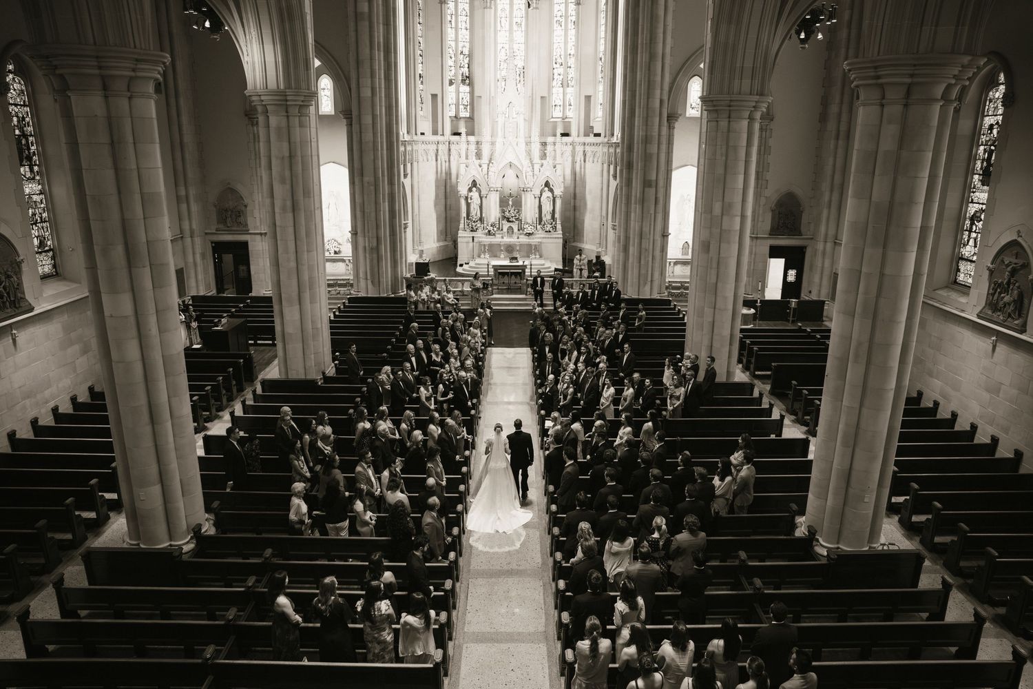 Black and white aerial view of a wedding ceremony in a grand cathedral with guests seated in wooden pews.