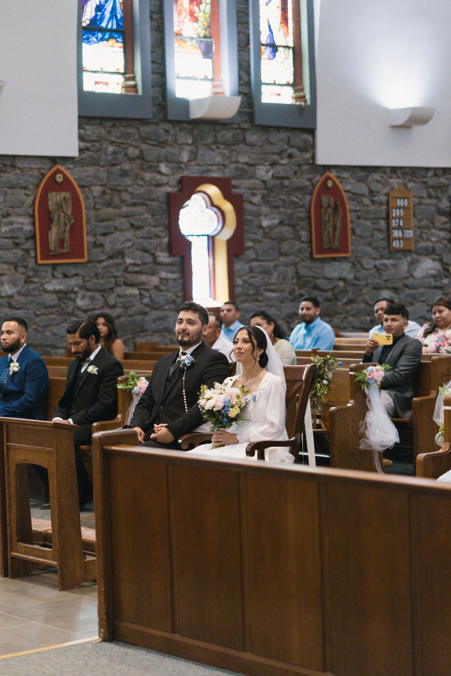 Newlywed couple sits together in church pews surrounded by wedding guests during ceremony.