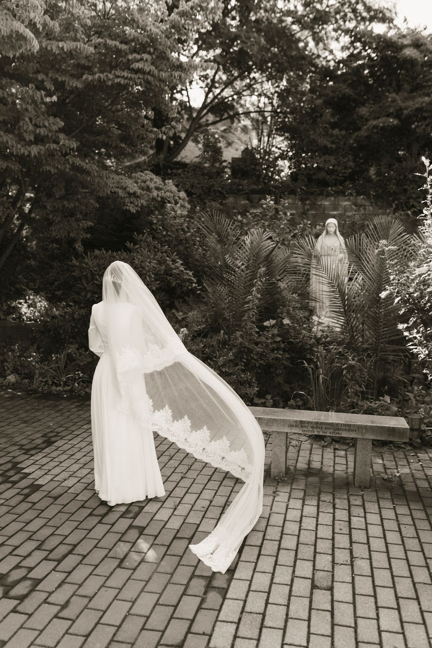 Bride in white gown with lace-trimmed veil walks toward garden statue in sepia-toned wedding photo.
