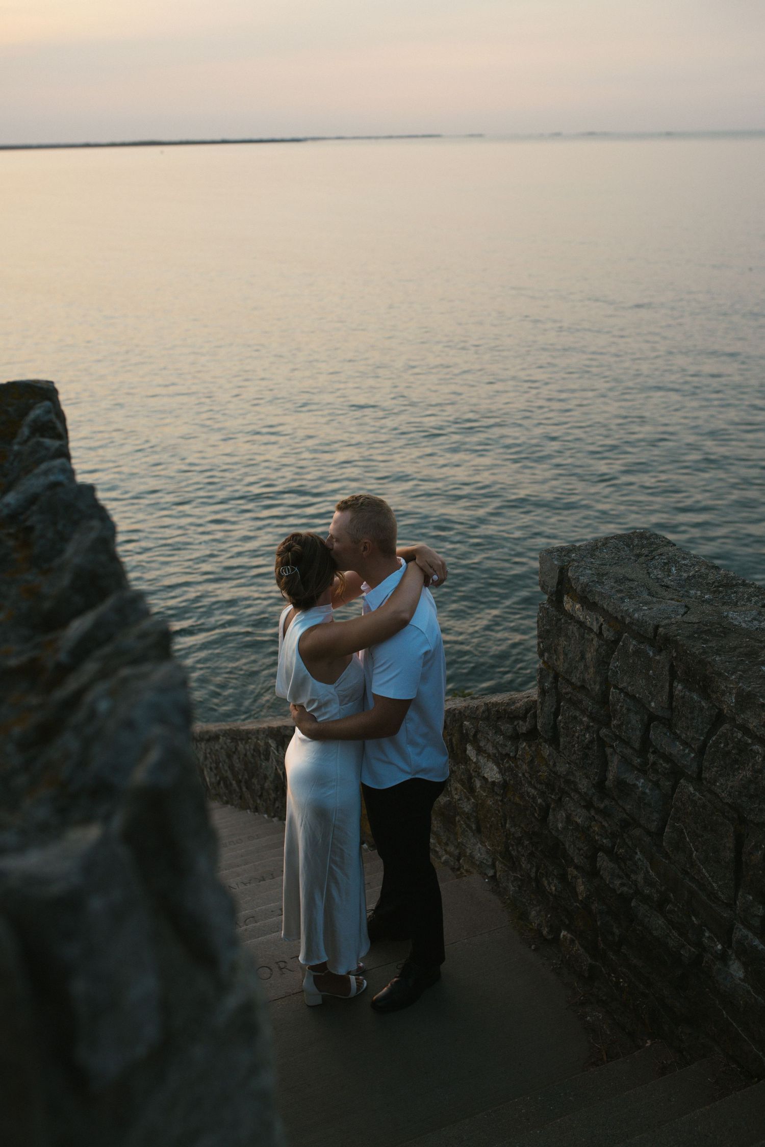Couple embracing and kissing at sunset by a stone wall overlooking calm ocean waters.
