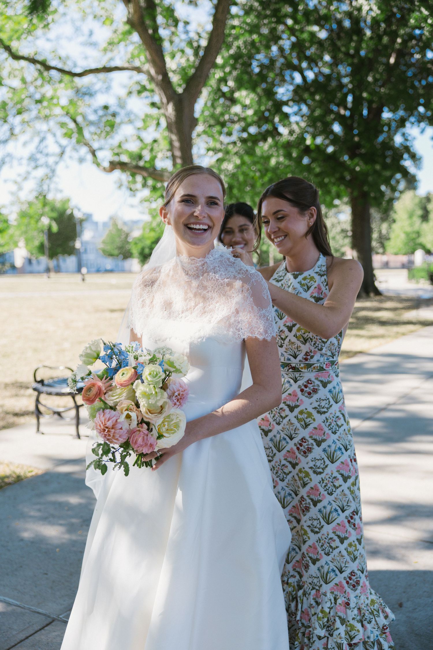 Laughing bride in white gown and veil holds pastel bouquet while bridesmaids adjust her dress outdoors.