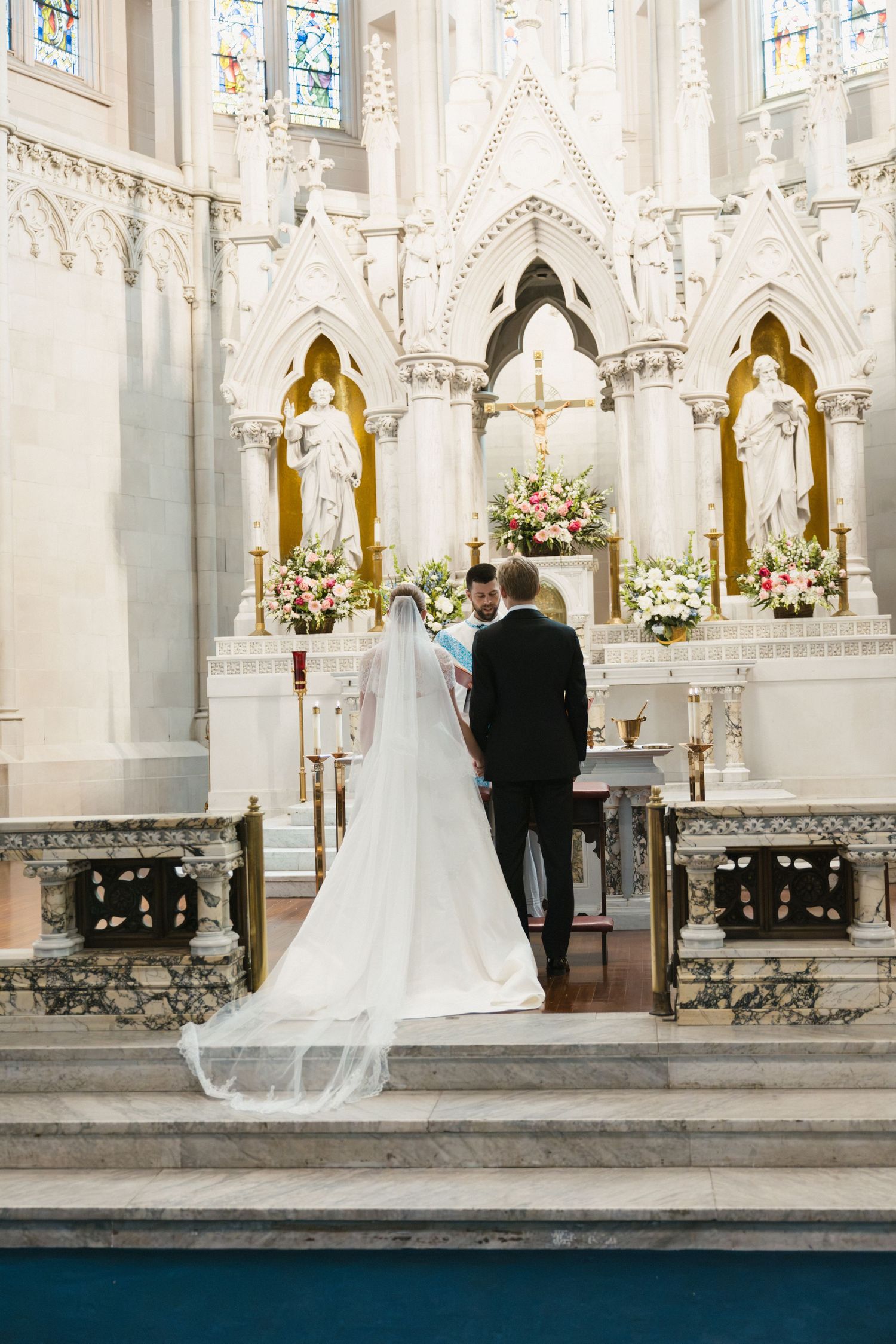 Bride in white gown and veil stands at ornate church altar with groom during wedding ceremony.