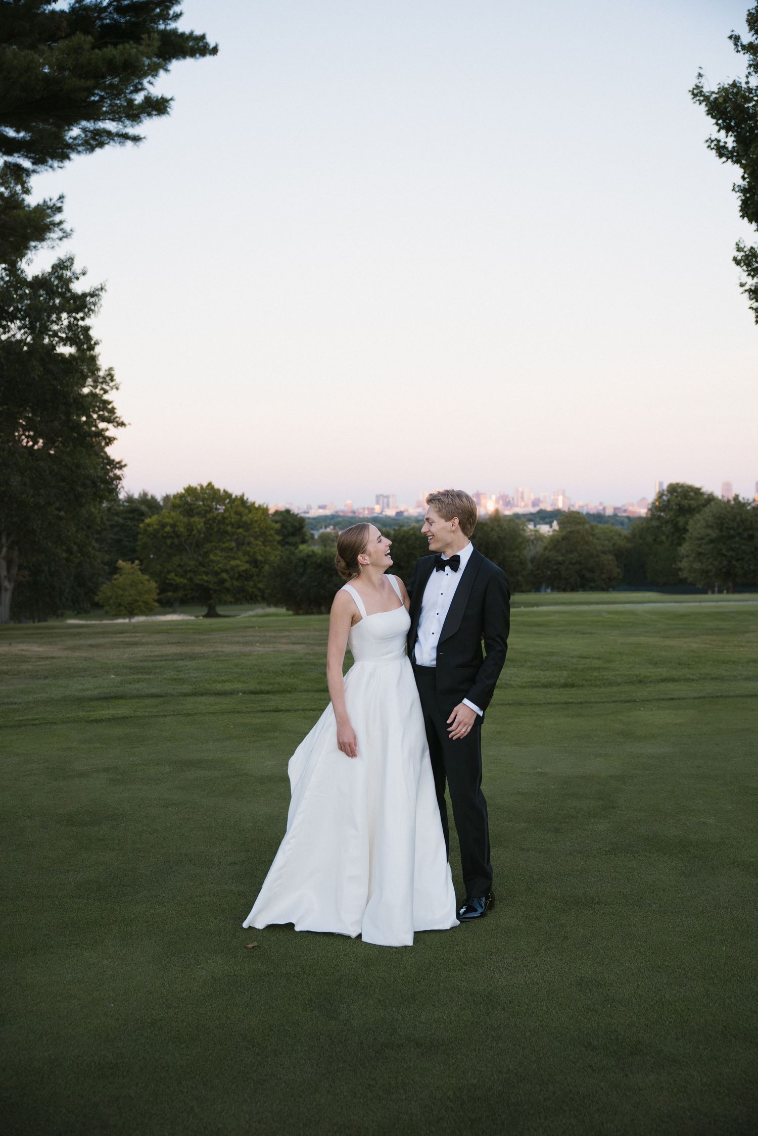 Bride and groom gazing at each other on a golf course at dusk with city skyline in background.