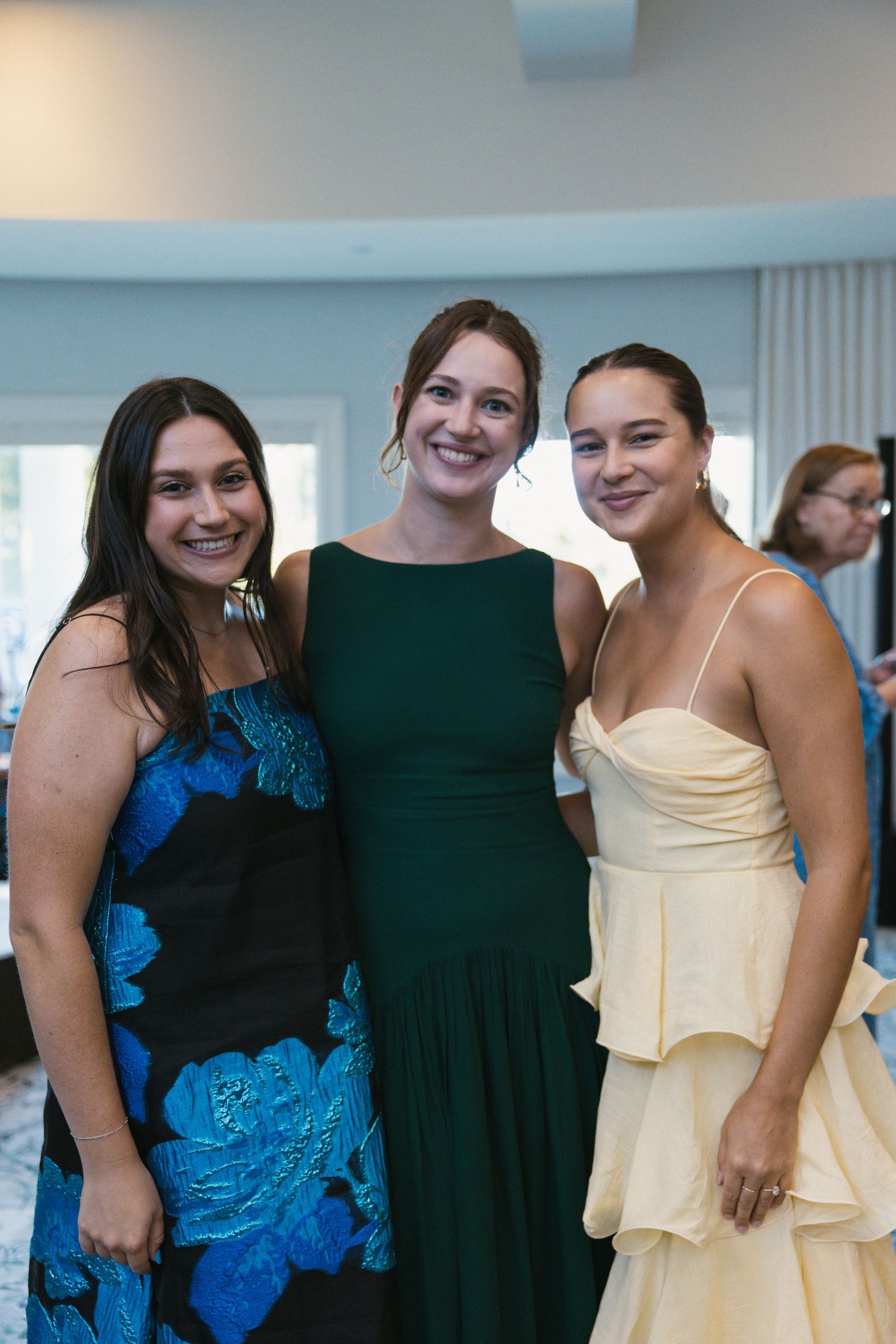 Three smiling women pose together at a formal event, wearing elegant dresses in blue floral, green, and cream.