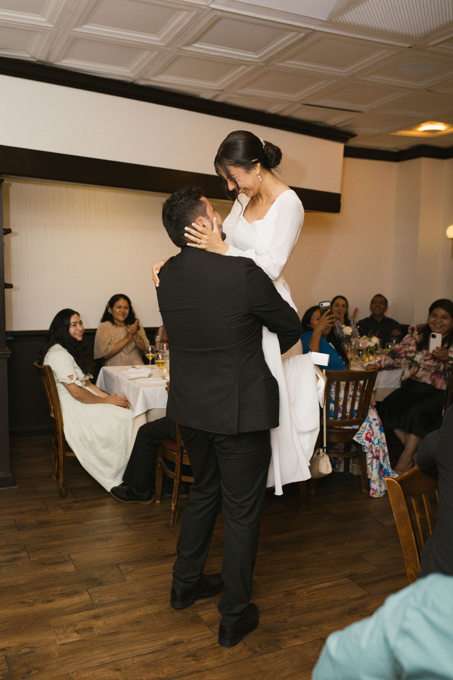 Groom lifts joyful bride during first dance at wedding reception while guests watch and celebrate.